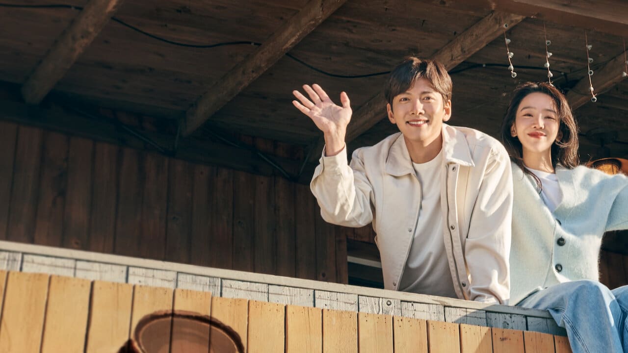 Một cặp đôi trẻ tươi cười vẫy tay chào từ ban công gỗ ấm cúng